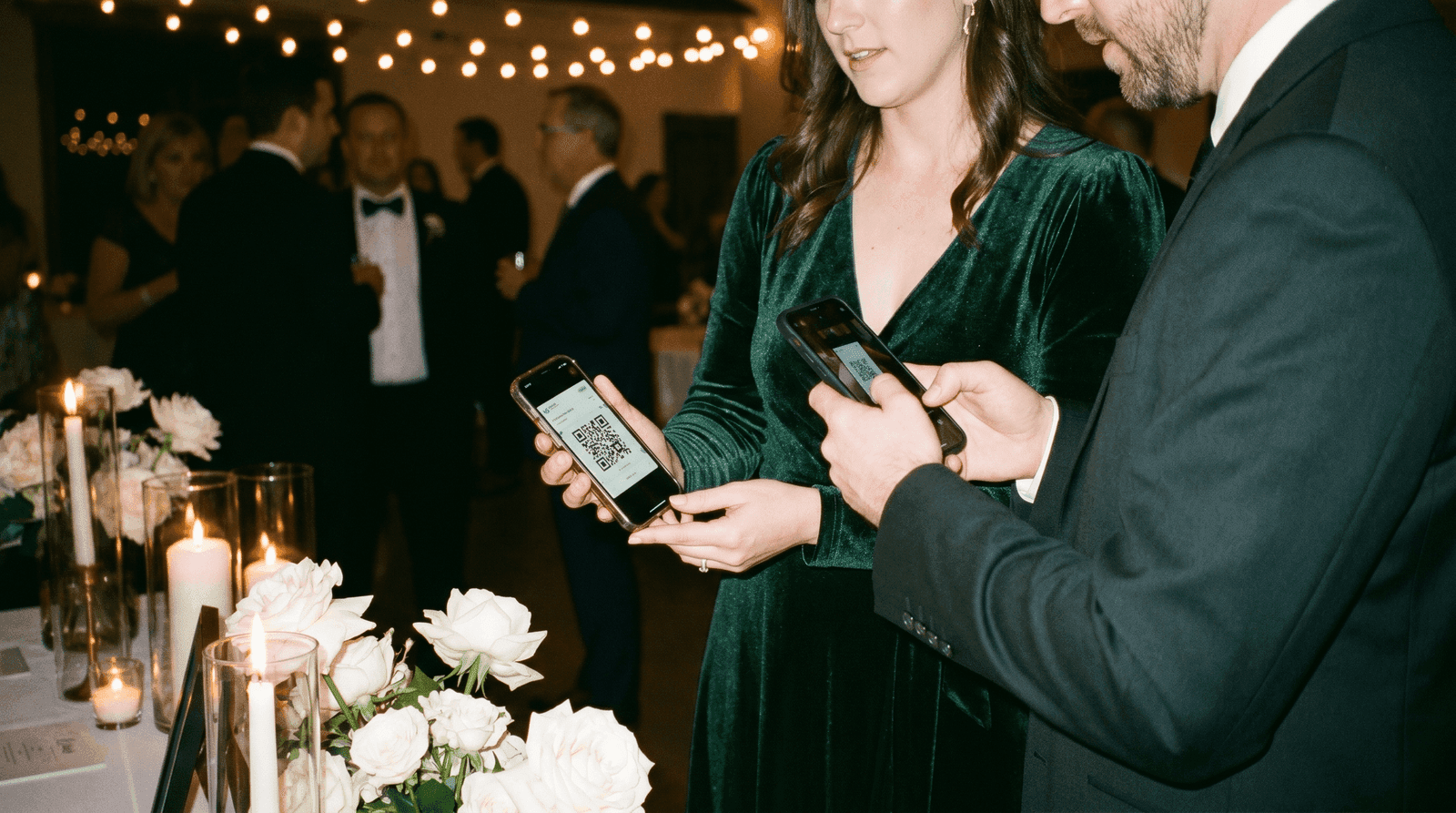 キャンドルの灯る周年パーティの受付で、緑のドレスの女性が提示するスマートフォンのQR招待状を、男性が自分のスマートフォンの背面カメラで読み取るシーン